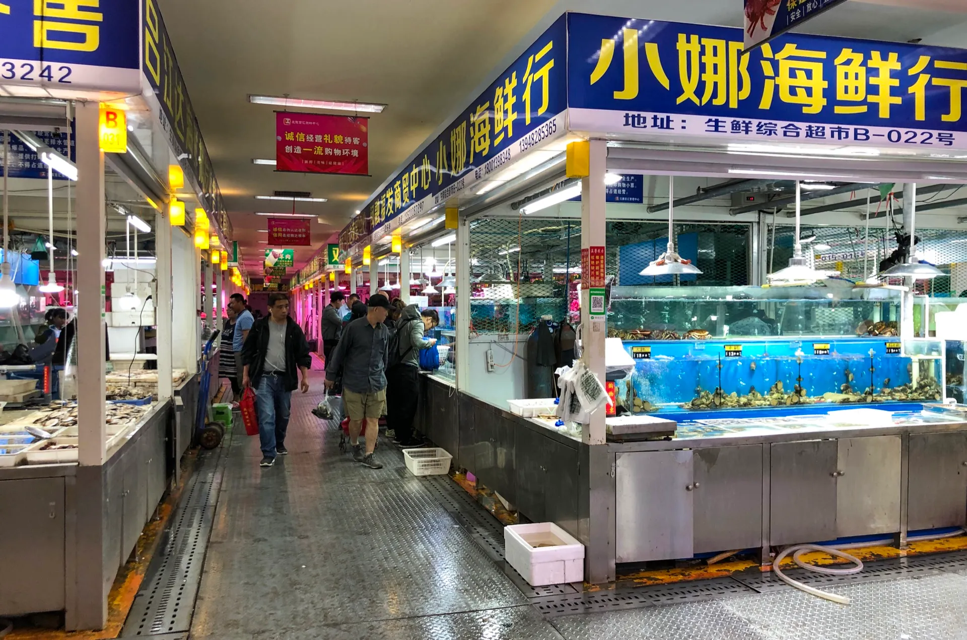 A bustling Dalian seafood market hall lined with live tanks and a red seafood-vendor sign