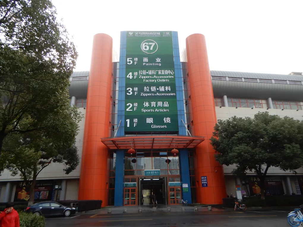 A directory-covered entrance at Yiwu International Trade City listing a different category of goods on each floor — glasses, sports articles, zippers, painting